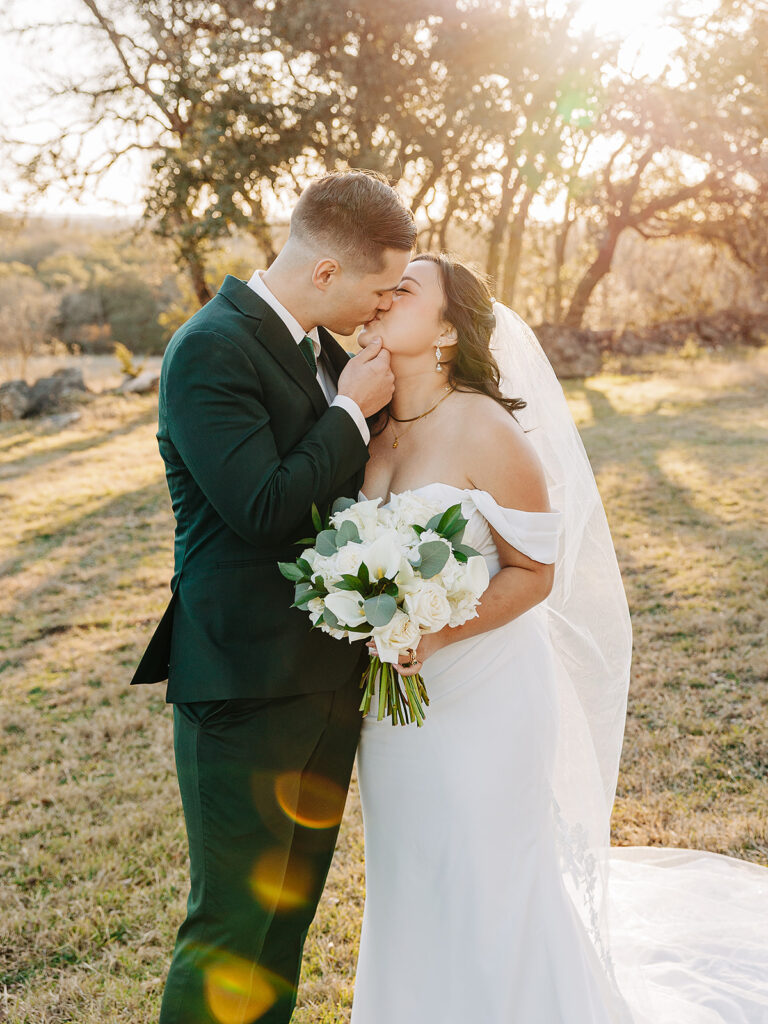 A bride and groom share a kiss outdoors at sunset. The bride wears an off-shoulder white dress, holds a bouquet of white flowers, and has a veil. The groom wears a dark suit. Sunlight filters through the trees behind them.