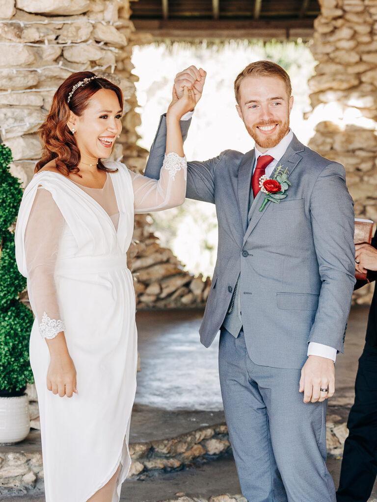 A bride in a white dress and a groom in a blue suit stand smiling, holding hands up in celebration, under a stone archway at their wedding ceremony.