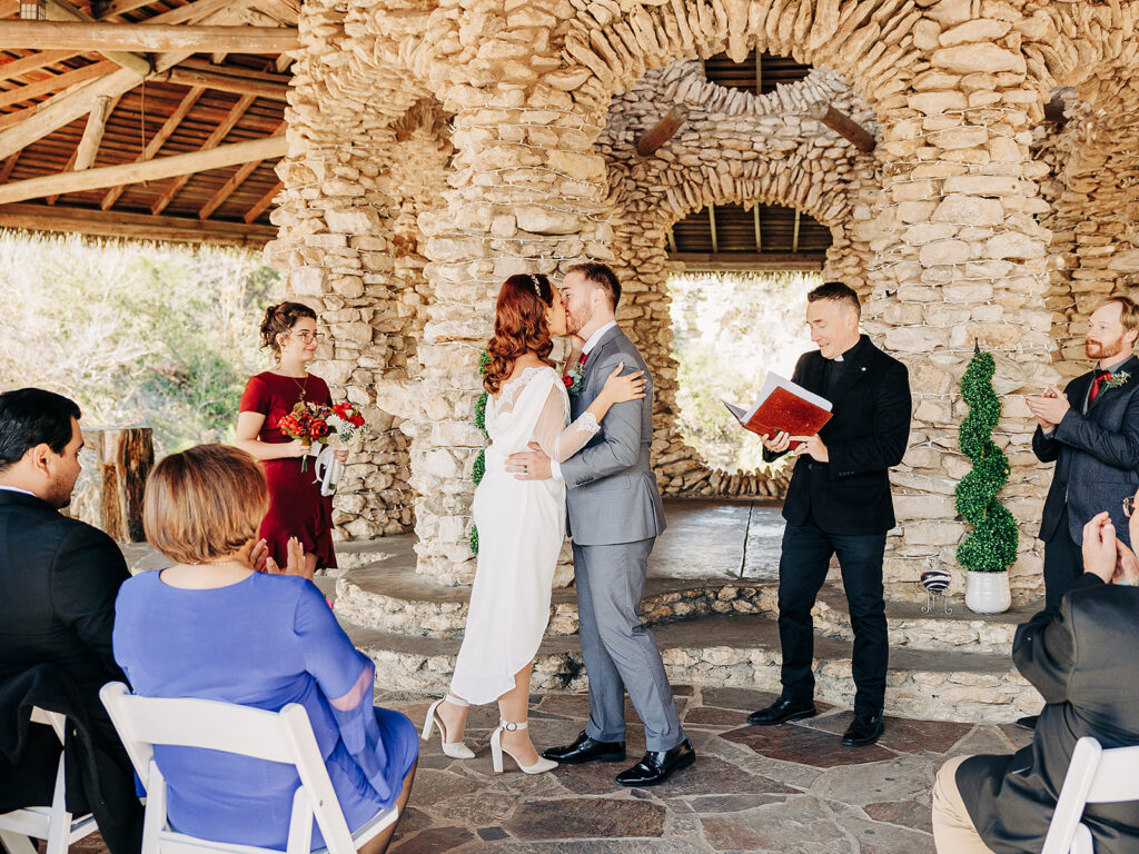 A bride and groom kiss at their wedding ceremony under a rustic stone pavilion, surrounded by clapping guests and attendants, with a celebrant holding a red book nearby.