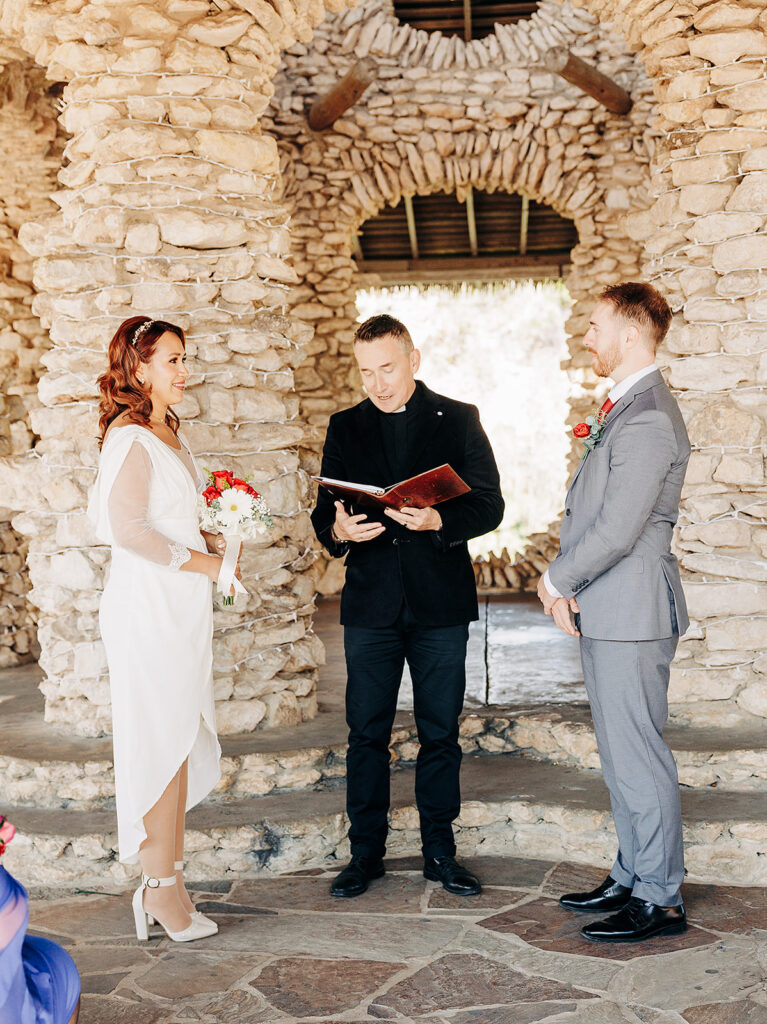 A bride in a white dress and a groom in a gray suit stand facing each other while an officiant reads from a book during a wedding ceremony in front of a rustic stone backdrop.