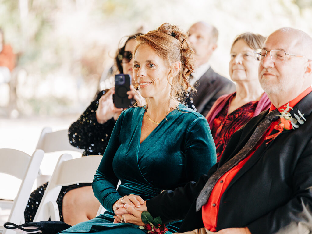 A woman in a teal dress sits holding hands with a man in a suit. They are seated among other people, all watching something off-camera at what appears to be a formal event or ceremony.