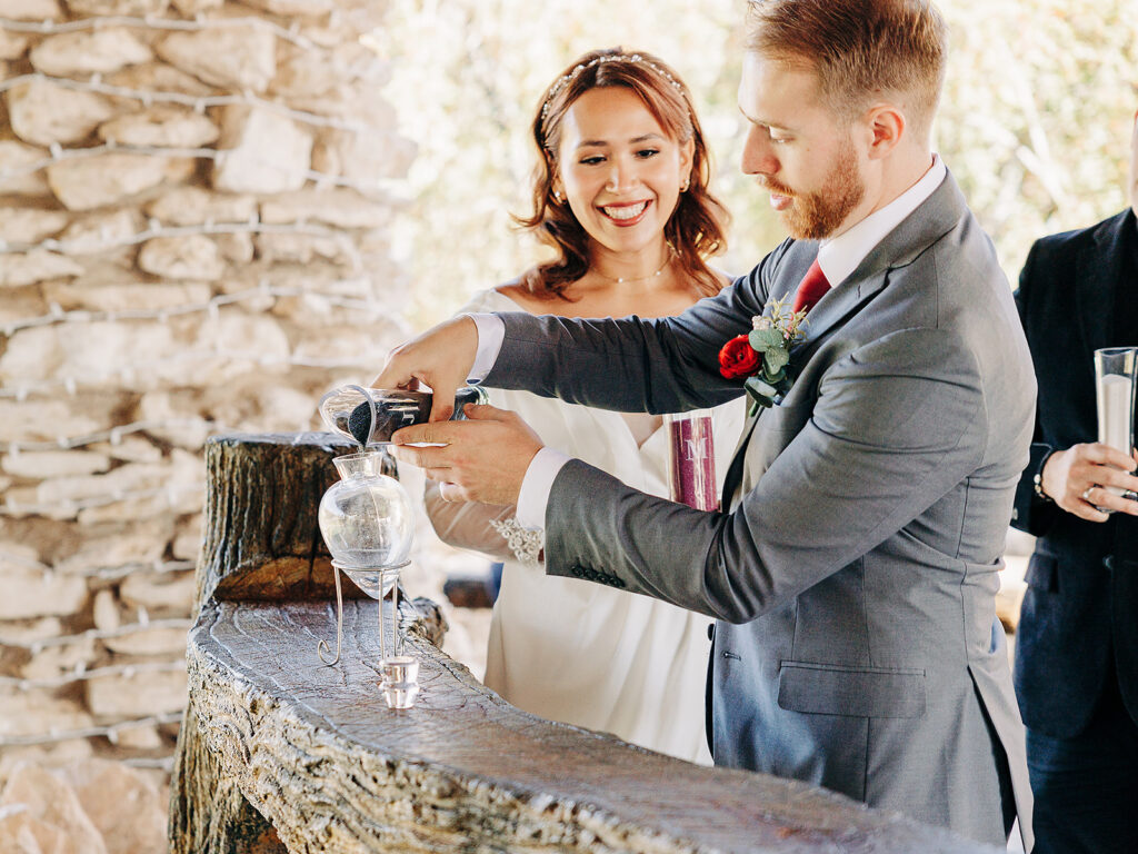 A bride and groom perform a sand ceremony together at their wedding. The bride smiles brightly while the groom pours sand into a glass vessel. They stand beside a rustic stone wall.