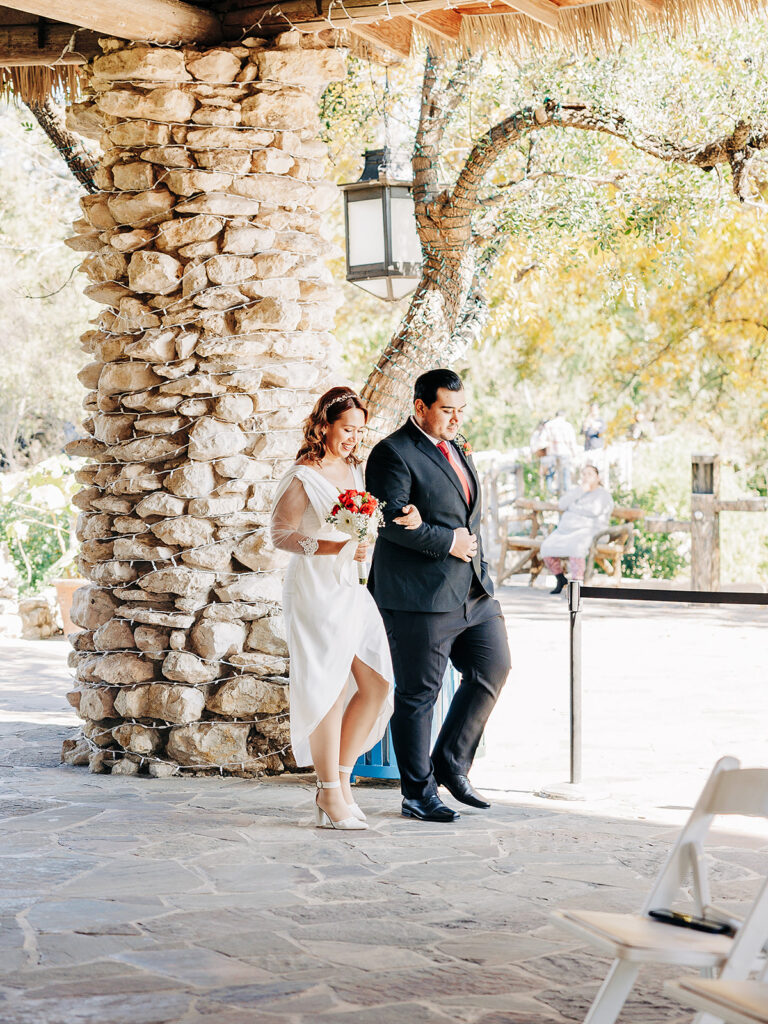 A woman in a white dress holding a bouquet walks arm-in-arm with a man in a black suit and red tie, outdoors near a stone pillar under a wooden canopy.