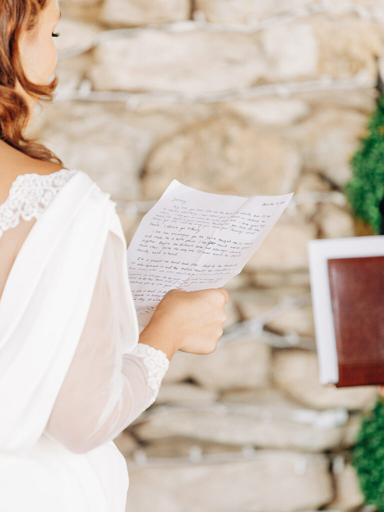 A bride in a white dress holds and reads a handwritten letter during a ceremony, with a blurred stone wall in the background.