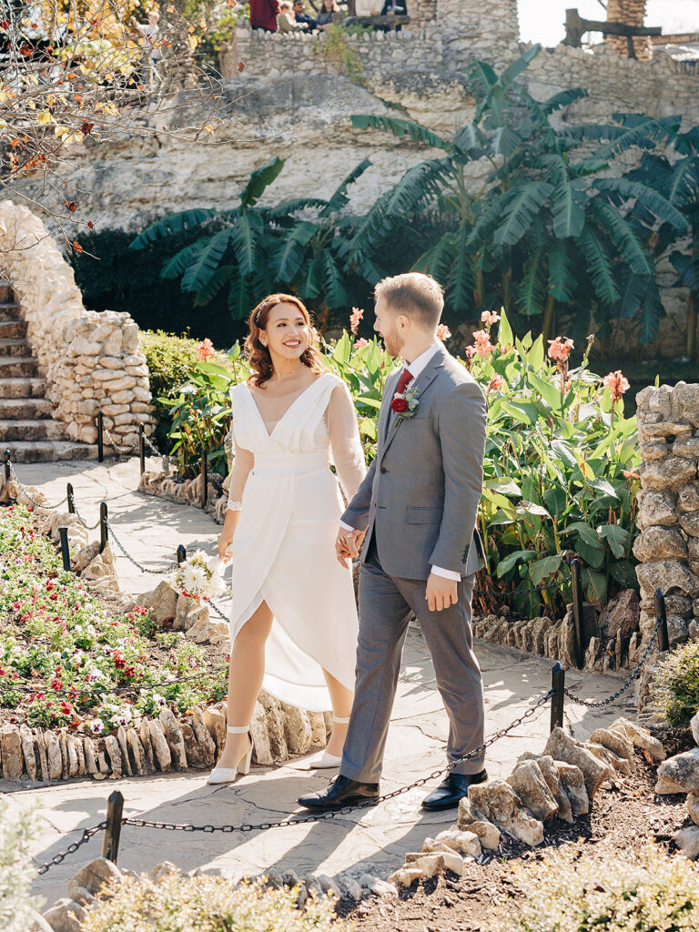 A bride in a white dress and a groom in a gray suit walk hand-in-hand on a garden path, surrounded by flowers, greenery, and stone walls, smiling at each other on a sunny day.