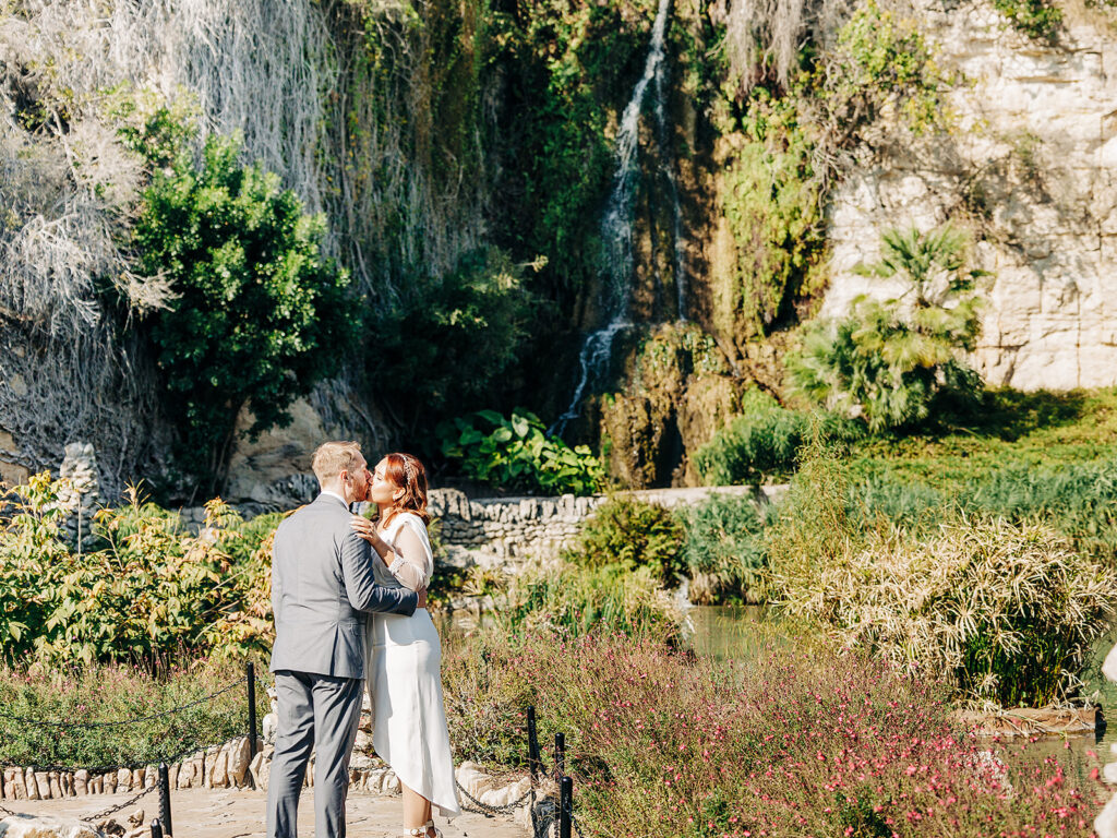 A couple in wedding attire embraces near lush greenery with a waterfall cascading down a rocky cliff in the background on a sunny day.