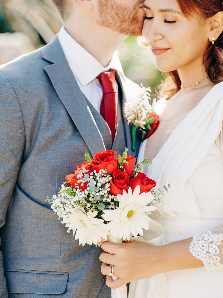 A bride in a white dress holding a bouquet of red roses and white daisies smiles as the groom in a grey suit and red tie gently kisses her forehead.