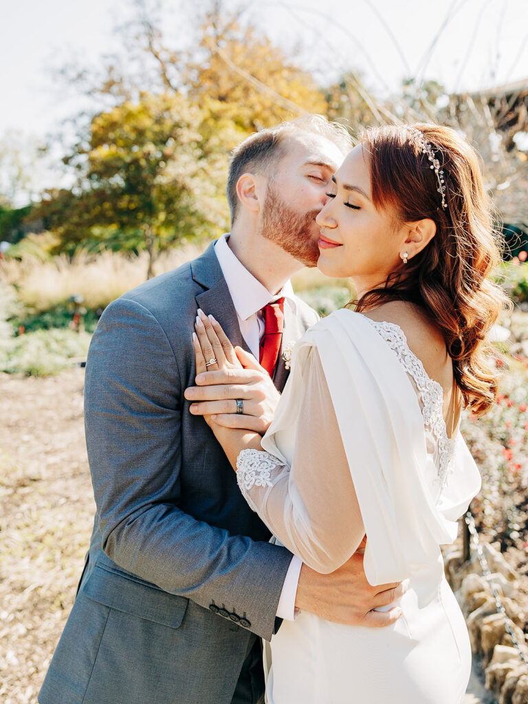 A groom in a gray suit kisses his bride on the cheek. The bride, wearing a white lace wedding dress with sheer sleeves, smiles with her eyes closed. They stand outdoors with greenery and sunlight in the background.