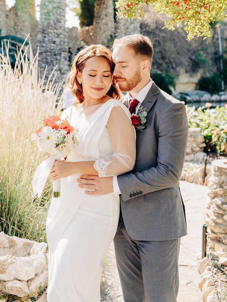 A bride in a white dress holding a bouquet stands closely with a groom in a gray suit. They embrace outdoors in a sunlit garden, both with peaceful, content expressions.
