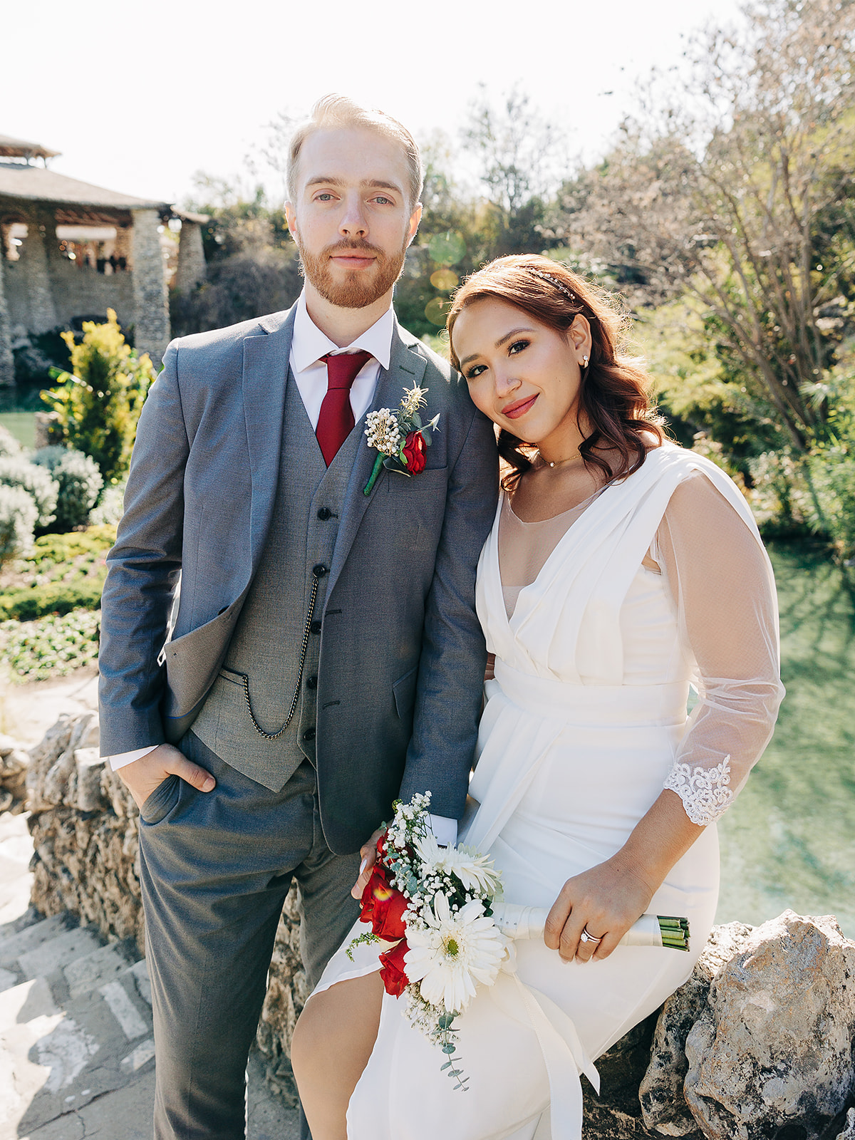 A bride in a white dress holding a bouquet and a groom in a gray suit with a red tie pose together outdoors by a pond, both looking at the camera on a sunny day.