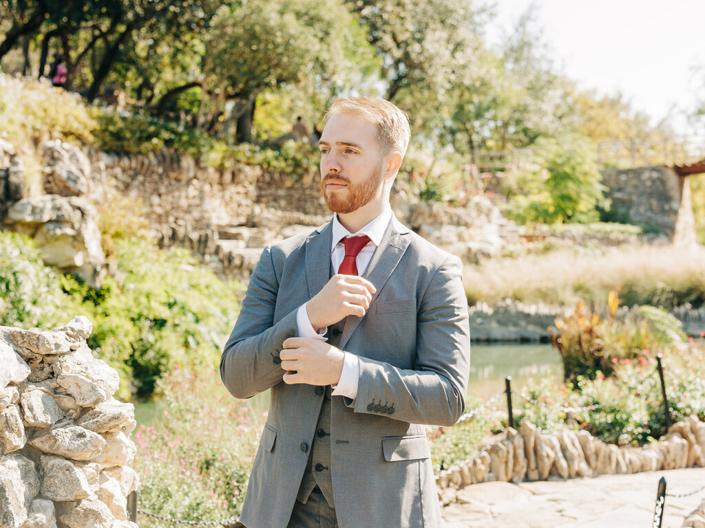 A man in a gray suit adjusts his cuff while standing outdoors near a stone wall, with greenery, flowers, and a pond in the background on a sunny day.