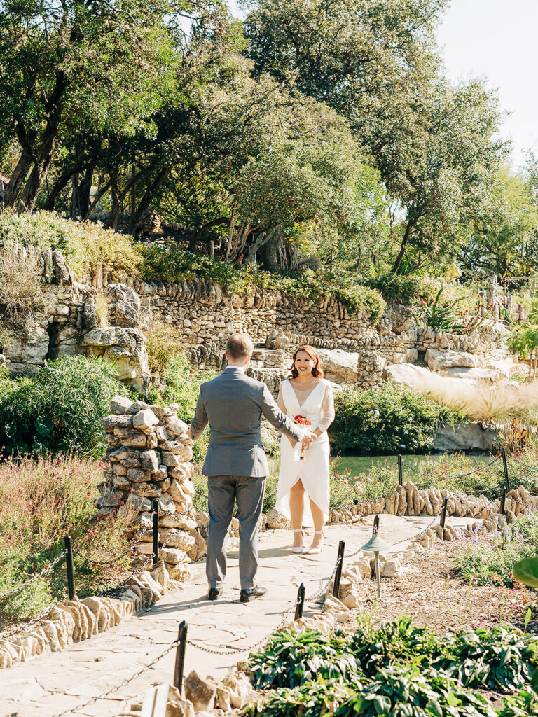 A man in a gray suit stands on a stone path, reaching out to a woman in a white dress holding flowers. They are surrounded by lush greenery and rock gardens on a sunny day.