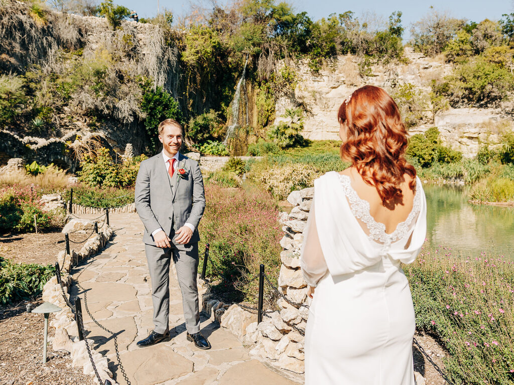 A groom in a gray suit smiles while facing a bride in a white dress with lace details. They stand on a stone path in a lush, sunny garden with greenery, a pond, and a waterfall in the background.