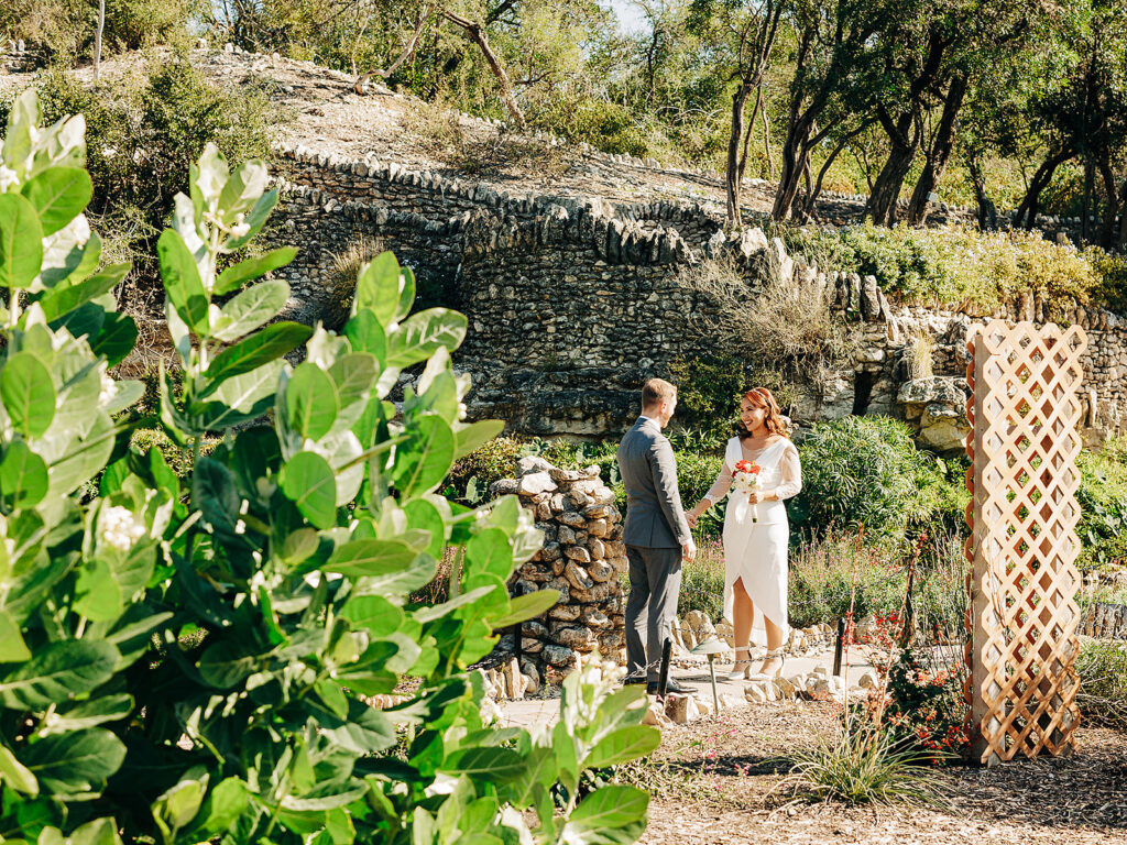 A couple stands facing each other in a garden, holding hands, surrounded by greenery, stone walls, and sunlight. The woman wears a white dress and the man wears a suit. A wooden trellis and lush plants are nearby.