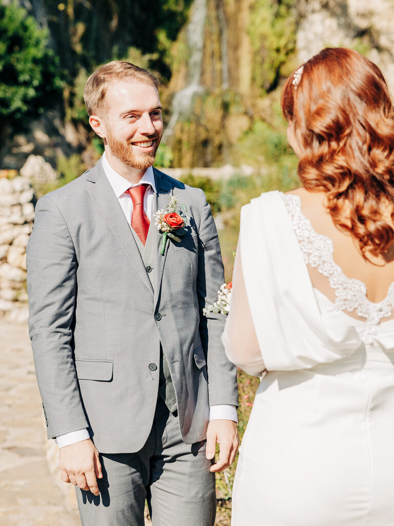 A groom in a gray suit smiles at a bride in a white dress with lace details, standing outdoors in sunlight with greenery and a waterfall in the background.