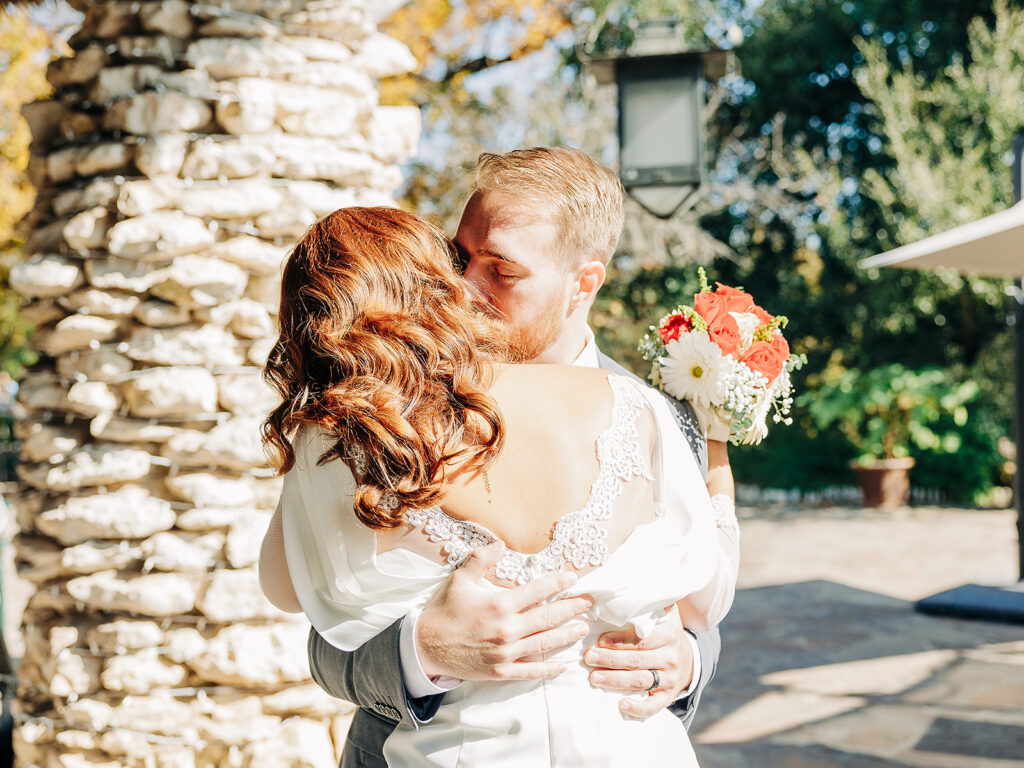 A bride and groom embrace and kiss outdoors in bright sunlight. The bride holds a bouquet of red and white flowers, while the groom wraps his arms around her. Stone columns and greenery are visible in the background.