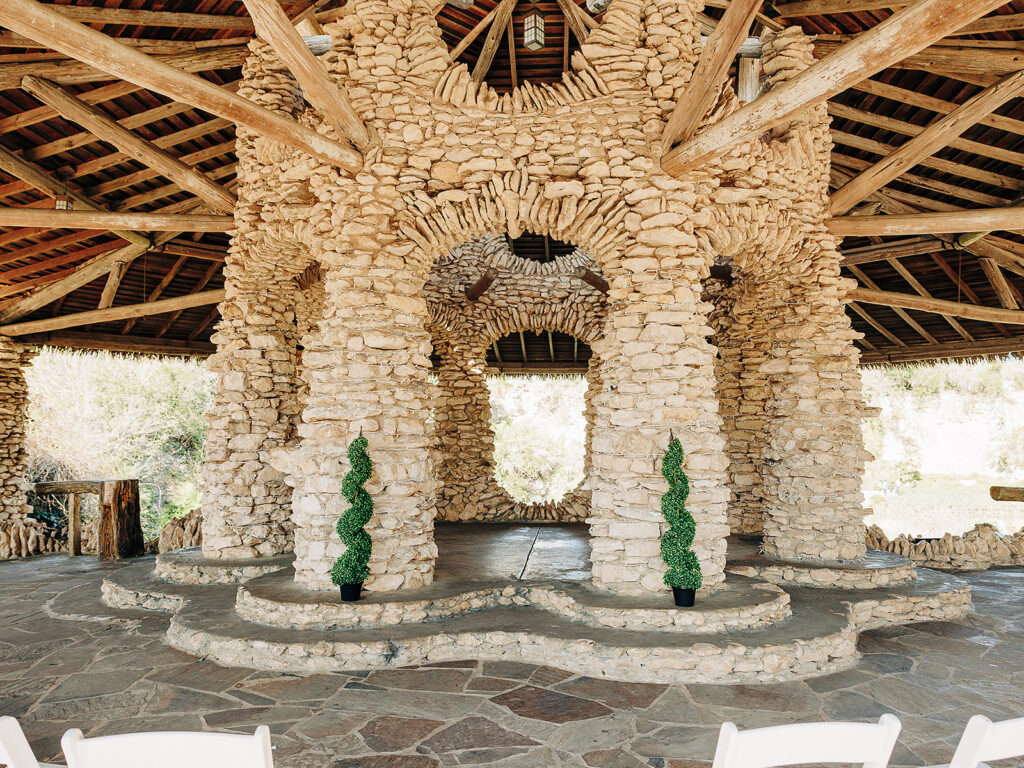 A stone pavilion with multiple round columns and arches, topped with a wooden roof. Two spiral topiary plants are placed at the entrance on a curved stone platform. Sunlight filters through the open structure.