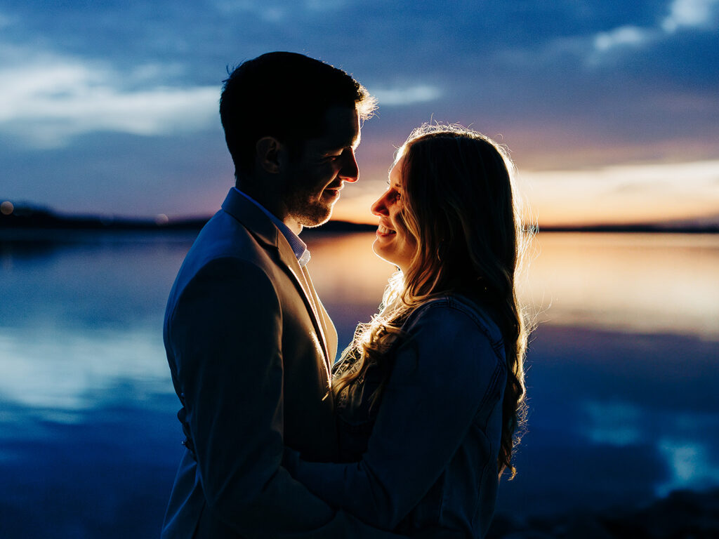 A couple embraces and smiles at each other by a calm lake at sunset, with the sky glowing in shades of blue and orange behind them.