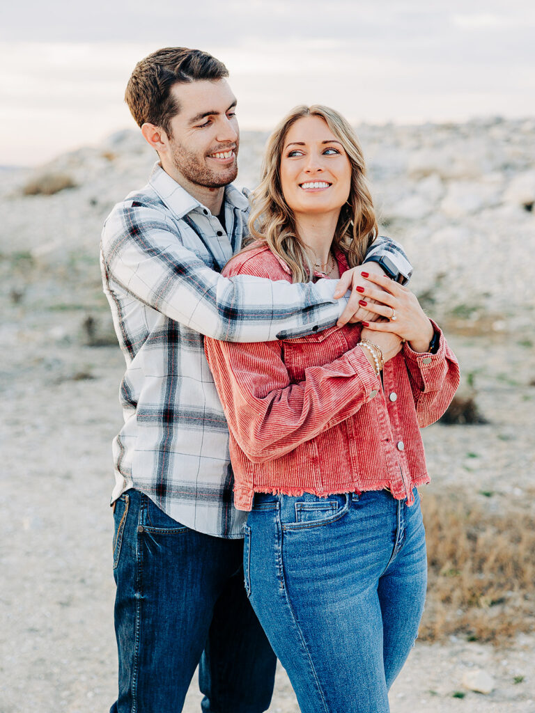 A young couple stands outdoors, smiling warmly. The man, in a plaid shirt and jeans, embraces the woman from behind. She wears a red jacket and blue jeans, holding his arms and looking off to the side. The background is rocky and natural.