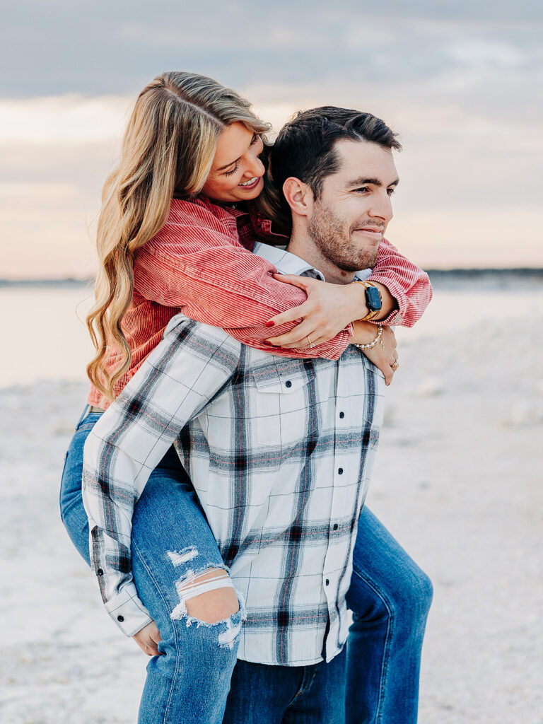 A woman in a red shirt and ripped jeans playfully hugs a man from behind as he gives her a piggyback ride on a beach, both smiling and looking happy. The background shows sand and a calm water with a soft, cloudy sky.