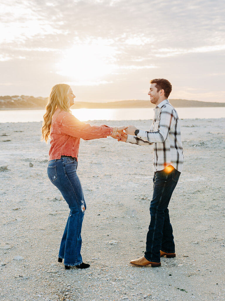 A couple stands on a sandy beach at sunset, holding hands and smiling at each other. The sun is low in the sky, casting a warm glow and creating lens flare on the serene landscape behind them.