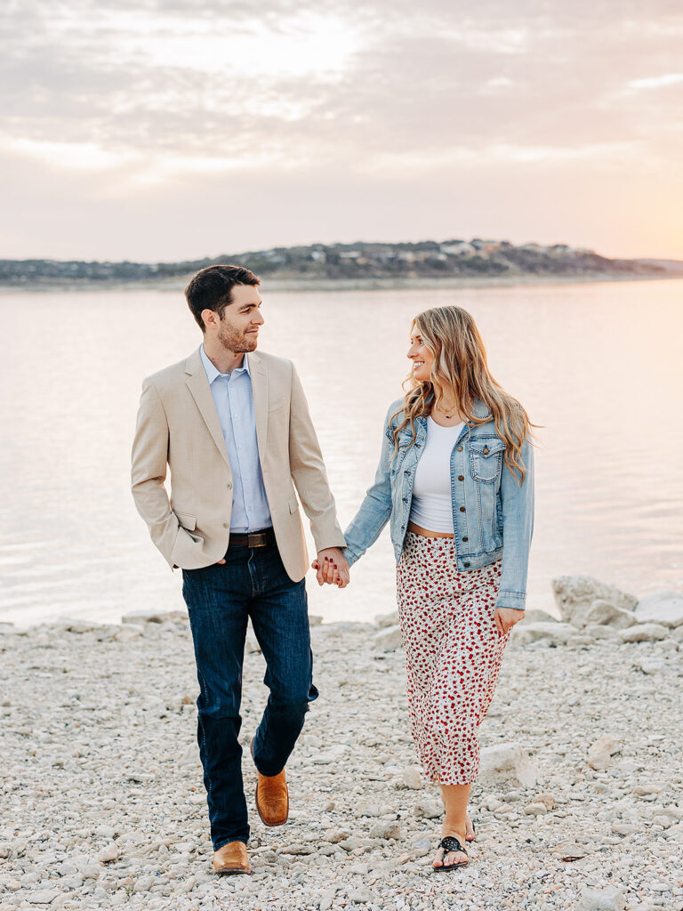 A man and woman hold hands and walk along a rocky lakeshore at sunset, smiling at each other. The man wears a beige blazer with jeans, and the woman wears a denim jacket and patterned skirt.