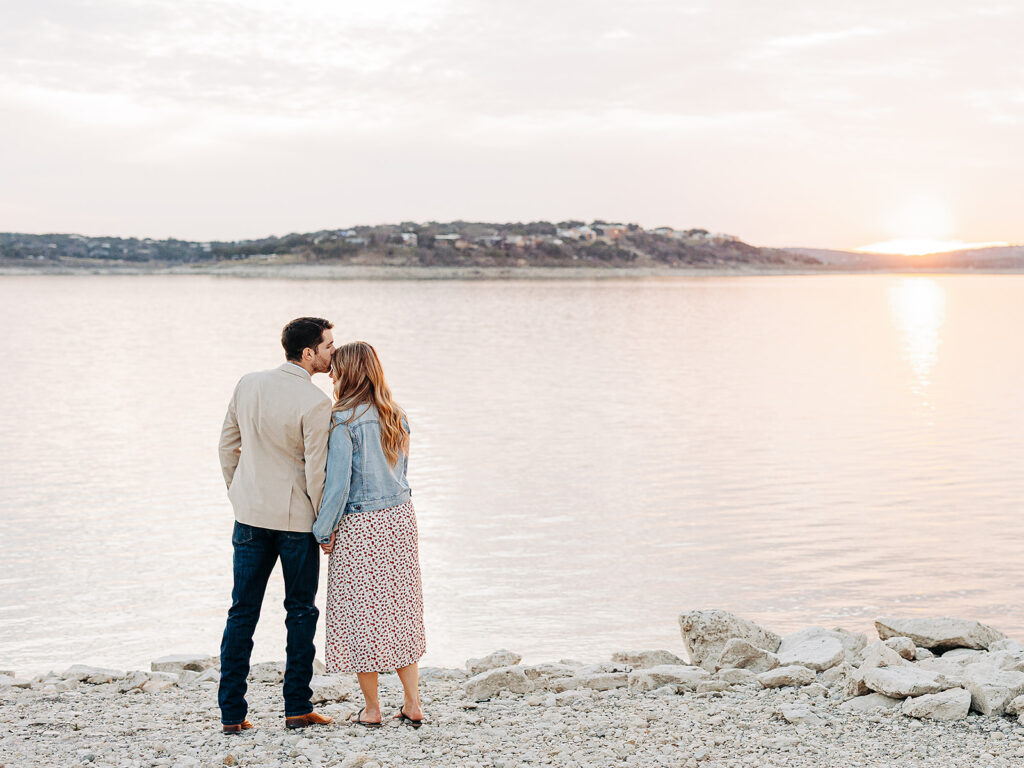 A couple stands on a rocky shore, facing a calm lake at sunset. The man kisses the woman on the forehead as they hold hands, with an island and distant houses visible across the water.