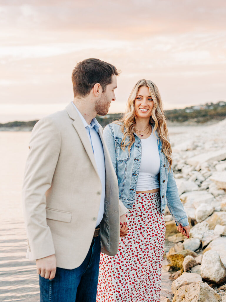 A man and woman walk hand in hand along a rocky shoreline at sunset. The man wears a beige blazer and jeans, while the woman wears a denim jacket, white top, and a floral skirt. They smile at each other.