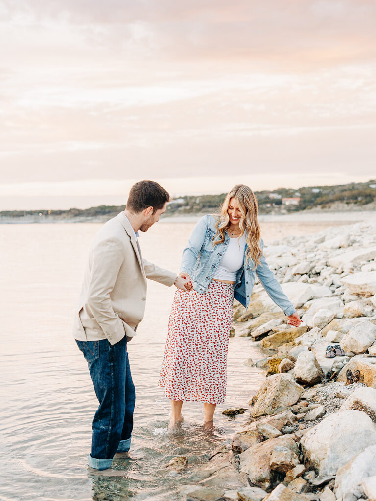 A man and woman hold hands and smile while walking along a rocky shoreline at sunset. The woman wears a floral skirt and denim jacket, and the man wears jeans and a beige blazer. Both have their feet in the shallow water.
