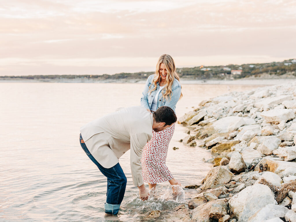 A couple stands by a rocky lakeshore at sunset; the man bends down to splash water near the womans feet as she laughs, wearing a patterned dress and denim jacket.