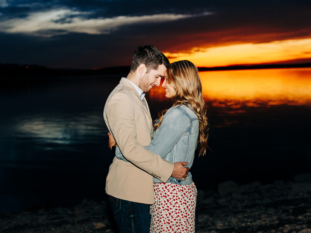 A couple stands close together by a calm lake at sunset, smiling and looking into each others eyes, with vivid orange and yellow hues reflecting on the water behind them.