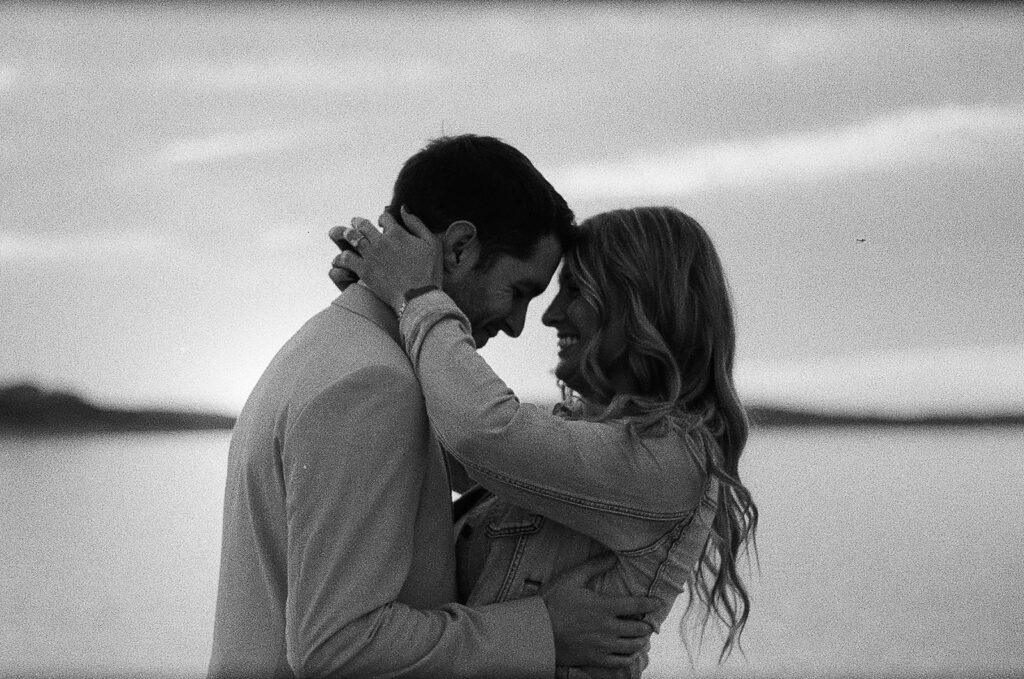 A couple embraces by a body of water at sunset, smiling and touching foreheads, captured in black and white.
