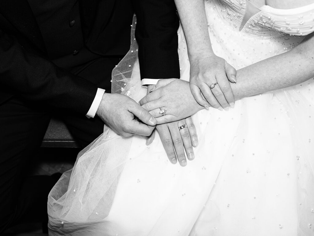 Black and white photo of a bride and groom in wedding attire sitting closely, holding hands with visible wedding rings, emphasizing the couple’s connection and commitment.