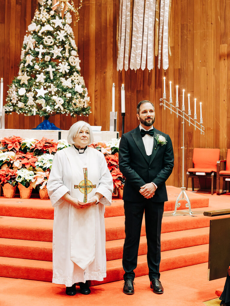 A man in a tuxedo stands beside a female officiant in white robes holding a cross, both on red-carpeted steps in a church decorated with a Christmas tree, poinsettias, candles, and wooden walls.
