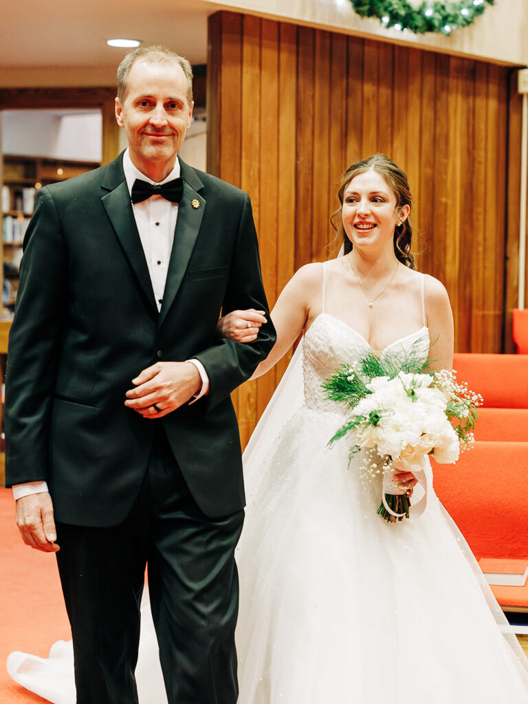 A bride in a white gown, holding a bouquet, walks arm in arm with a man in a black tuxedo down a red-carpeted aisle, both smiling inside a warmly lit indoor venue.