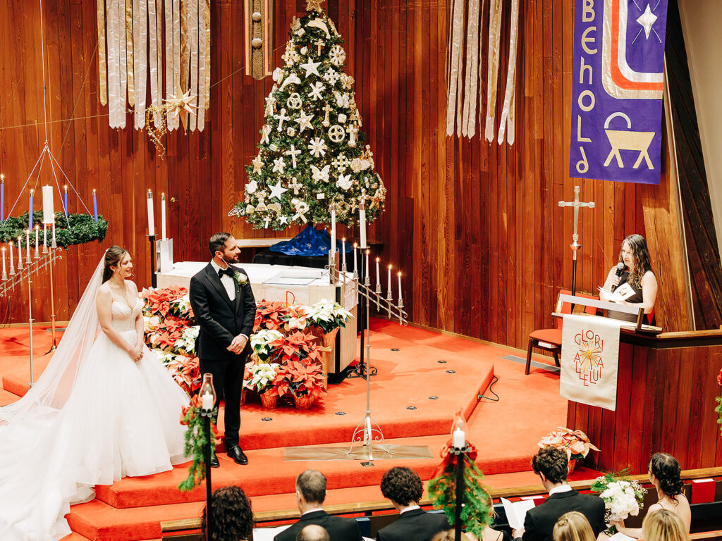 A bride and groom stand at the altar in a decorated church with red carpeting, a Christmas tree, poinsettias, and candles, while a woman at a podium addresses them. Guests are seated in the foreground.