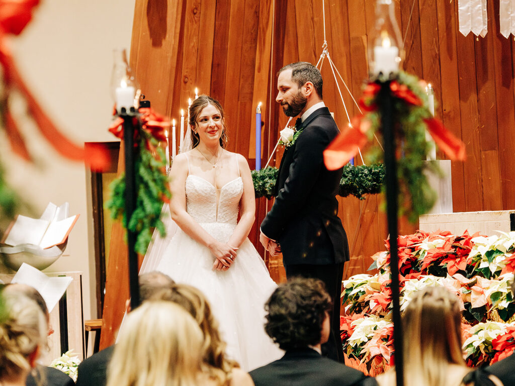 A bride and groom stand at the altar during a wedding ceremony, facing each other and smiling. Guests are seated in the foreground, and festive decorations and poinsettias adorn the scene.