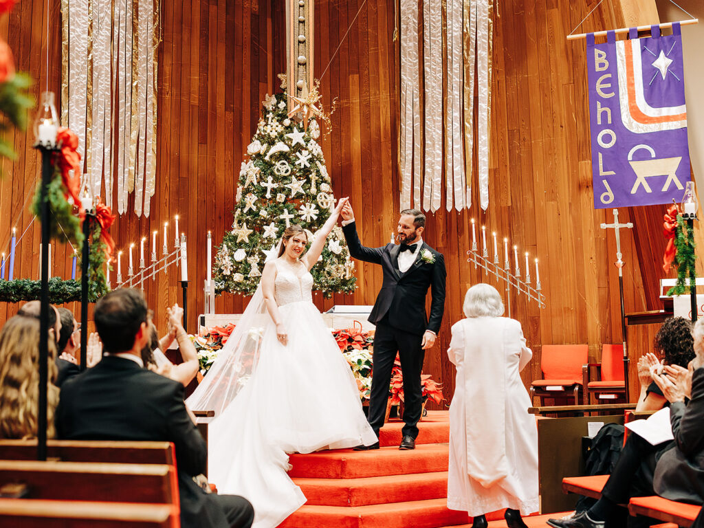 A newly married couple stands at the altar holding hands and smiling as guests applaud in a church decorated for Christmas, with a large tree, candles, and festive banners behind them.