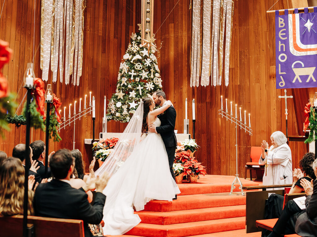 A bride and groom kiss at the altar in a decorated church with a Christmas tree, candles, and guests clapping. The scene is festive with red and gold accents and a large “BEHOLD” banner in the background.
