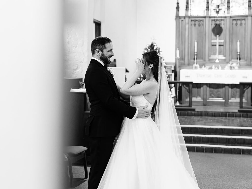 A bride and groom stand facing each other in a church, smiling and emotional. The bride wipes her eyes, wearing a long veil and white dress, while the groom holds her gently. The altar and cross are visible in the background.