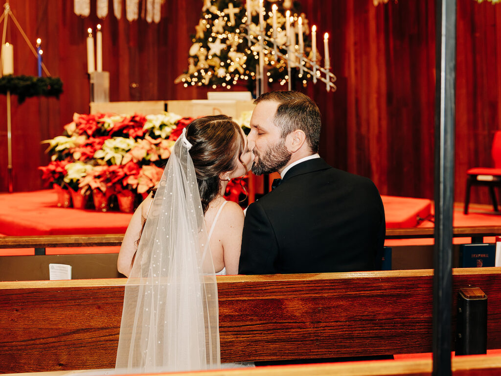 A bride and groom sit on a wooden pew, sharing a kiss in a church decorated with red flowers, candles, and a lit Christmas tree in the background. The bride wears a veil and the groom is in a suit.