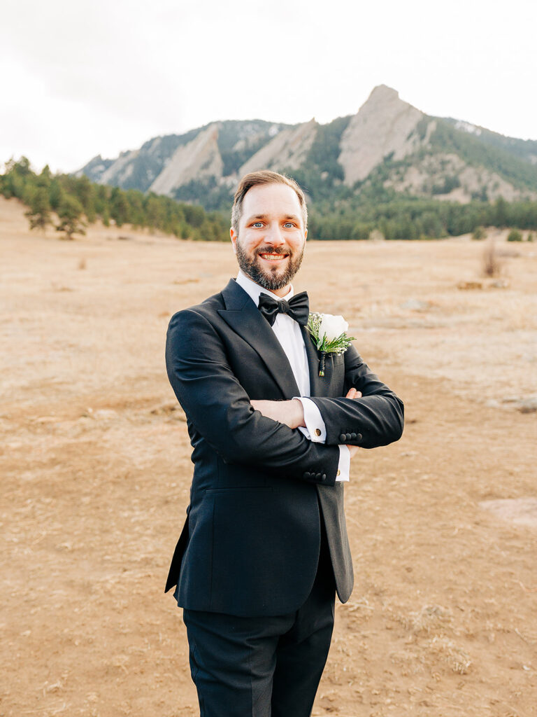 A man in a black tuxedo with a bow tie and boutonniere stands smiling with arms crossed in a dry, open field, with mountains and trees in the background.