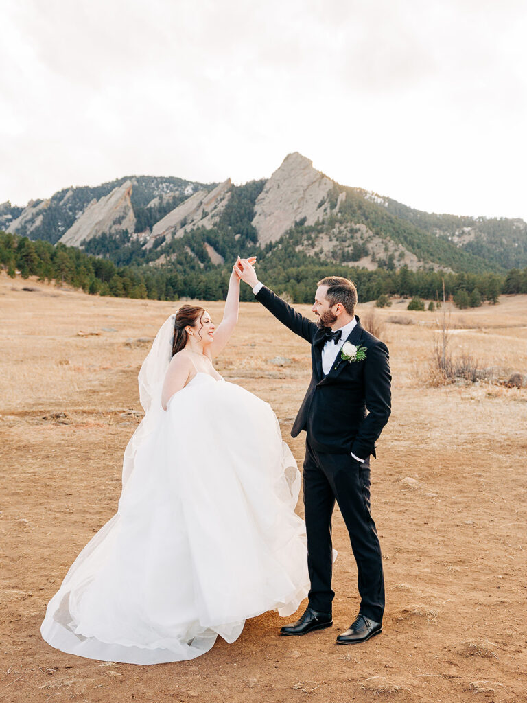 A bride in a white gown and a groom in a black suit dance outdoors on a dirt field, with mountains and pine trees in the background under a cloudy sky.