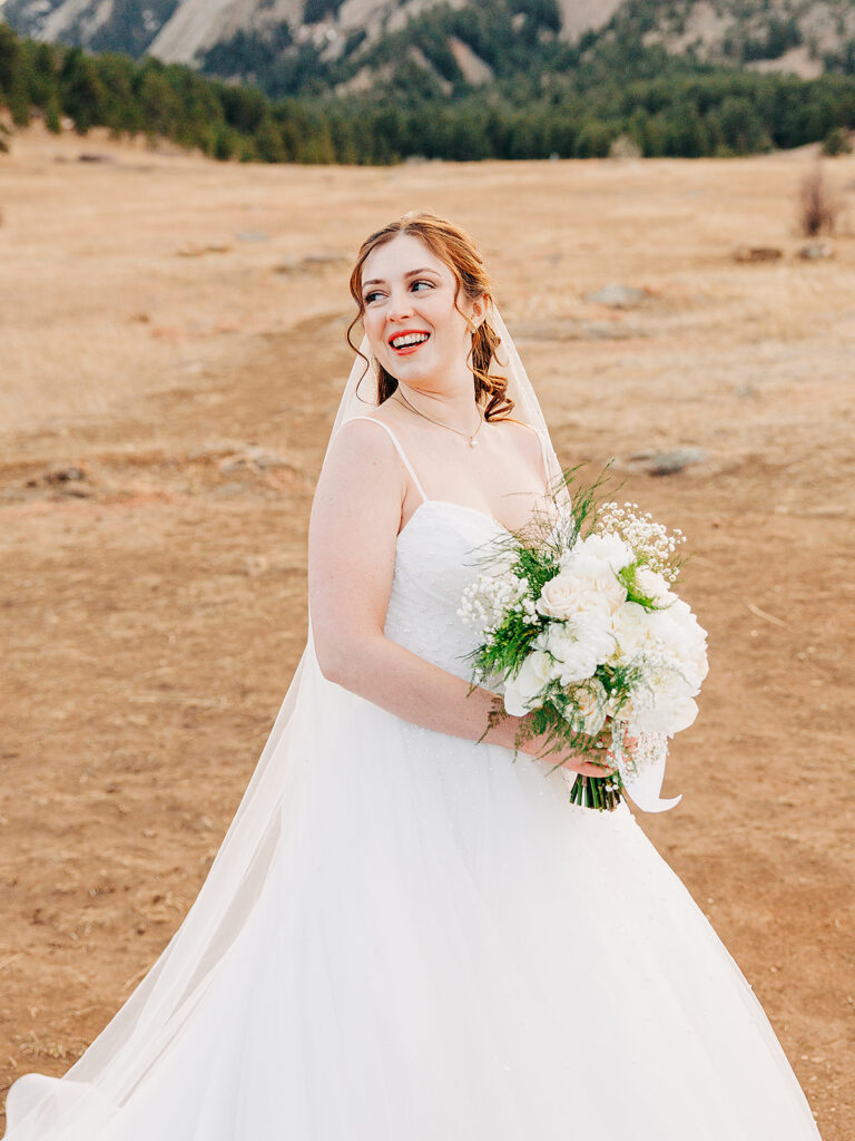 A smiling bride in a white wedding dress holding a bouquet of white flowers stands outdoors on a dry, grassy field with mountains and trees in the background.