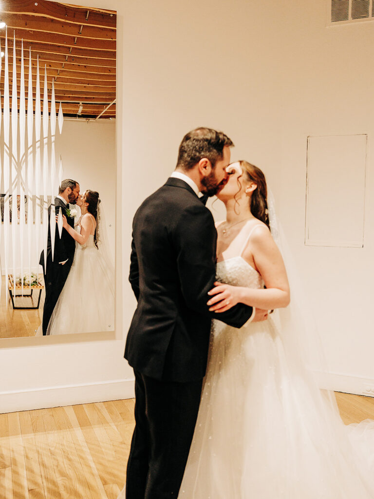 A bride and groom kiss indoors, dressed in wedding attire. Their reflection is visible in a large wall mirror behind them, adding depth to the intimate moment. The room has light walls, wooden floors, and modern decor.