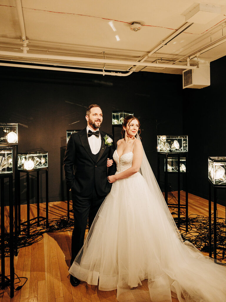 A bride in a white gown and veil stands smiling beside a groom in a black tuxedo. They are indoors, surrounded by glowing glass light fixtures on stands, on a wooden floor with a dark background.