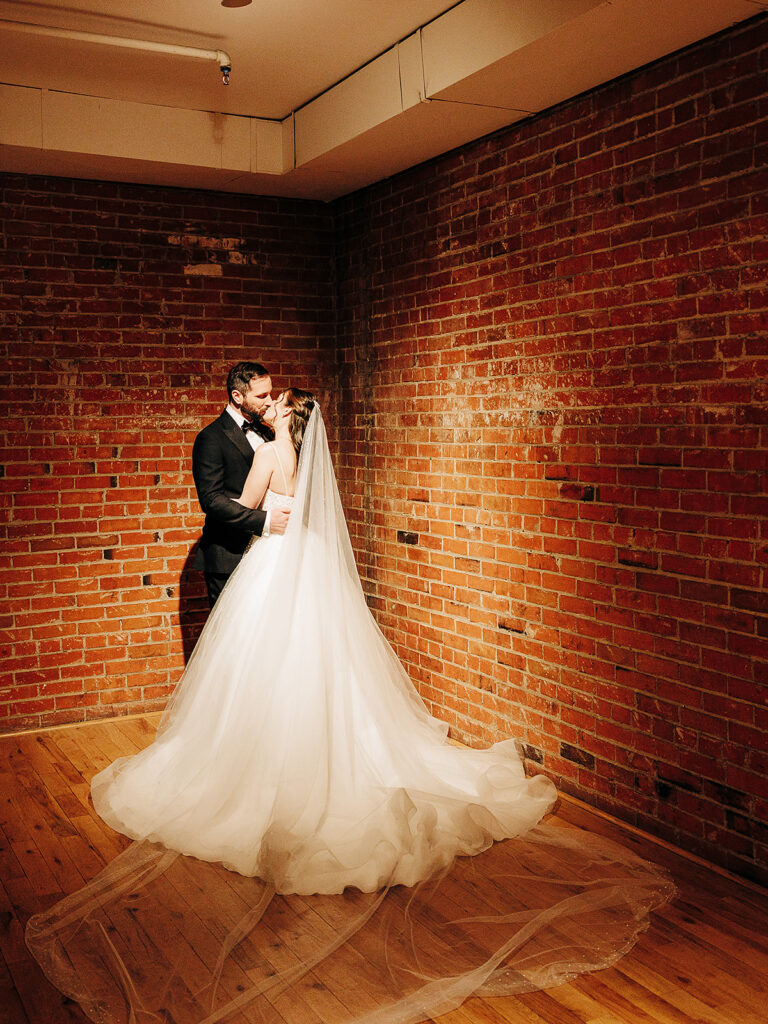 A bride and groom embrace and share a kiss in a warmly lit room with brick walls and a wooden floor. The bride wears a long, flowing white gown with a train and veil.