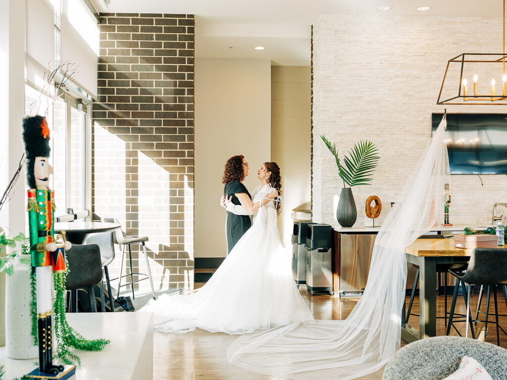 A bride in a white gown and long veil stands smiling and holding hands with a woman in black in a sunlit modern room, with a nutcracker decoration and plants nearby.