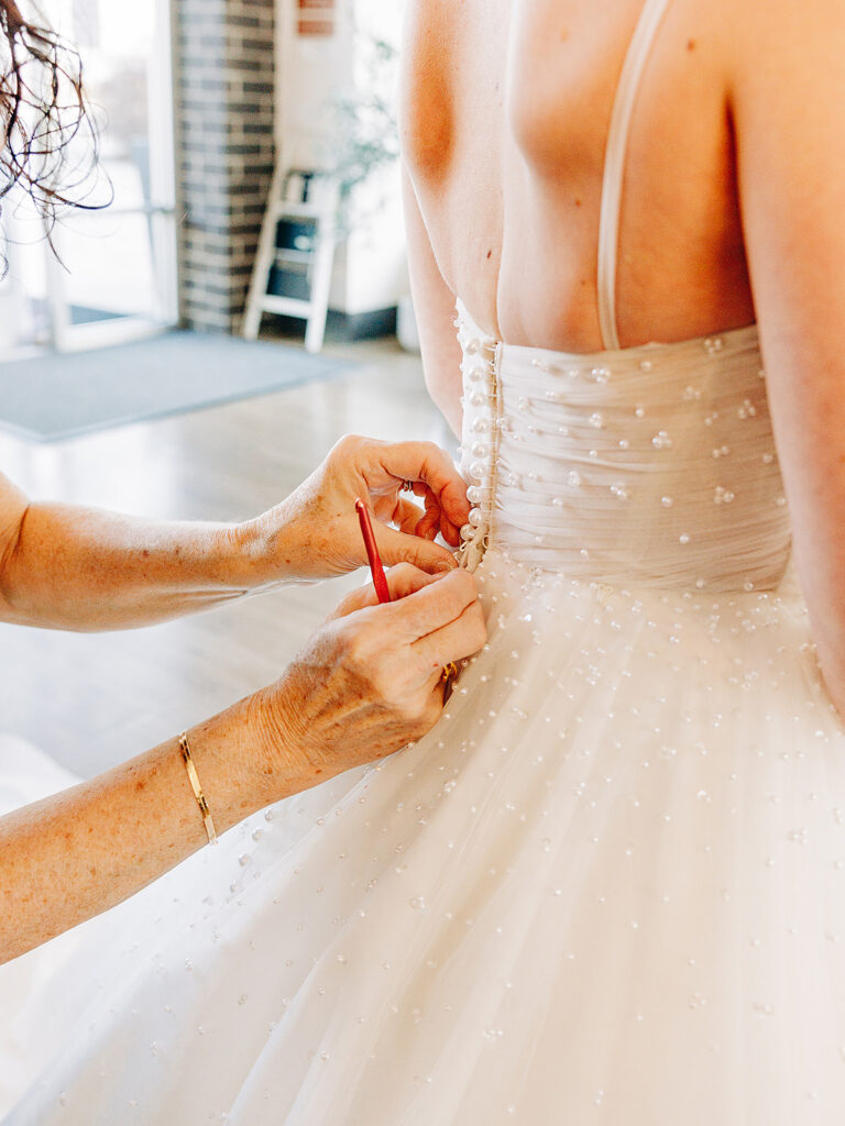A close-up of a woman fastening the buttons on the back of a bride’s white, beaded wedding dress, with the bride’s bare shoulders and the helpers hands visible.