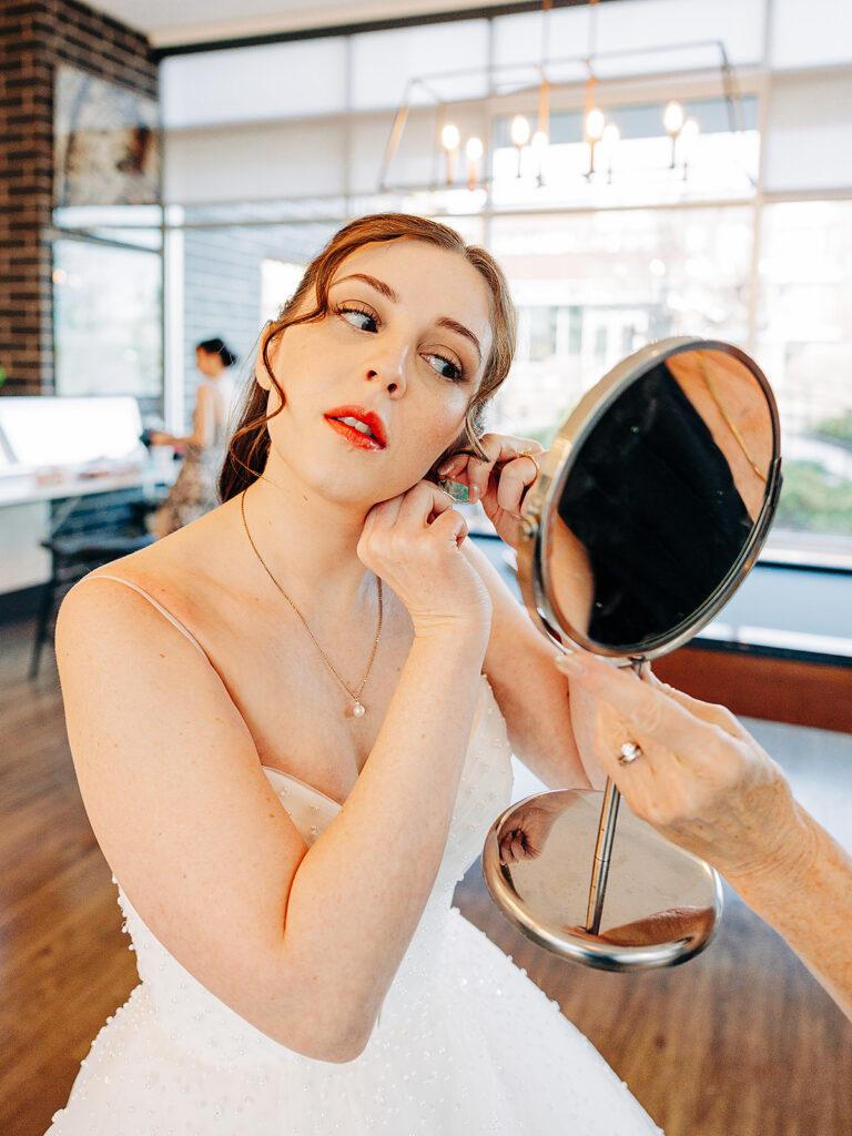 A bride in a white dress adjusts her earring while looking into a handheld mirror. She has red lipstick and her hair styled up. The background shows a bright room with large windows and another person in the distance.